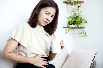 Asian middle-aged woman holding her stomach in pain after eating instant noodles. Mature female suffering from indigestion, stomachache, or acid reflux while sitting on a sofa in the living room