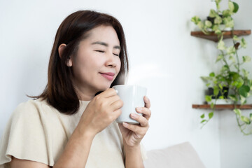 Peaceful Asian woman enjoying a hot cup of coffee at home. Relaxed mature lady closed eyes while smelling aroma of tea in bright room, representing morning routine, mindfulness, and self-care