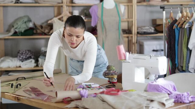 Young female seamstress drawing pattern on sheet of paper using curves while sewing dress at workplace in modern fashion sewing workshop