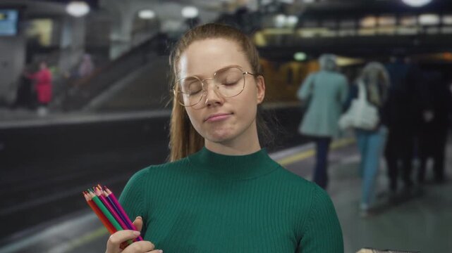 Young woman with eyeglasses holds colored pencils and sketchbook in train station building; curiosity.
