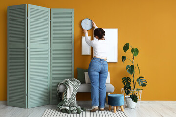 Young woman hanging clock on orange wall in room, back view