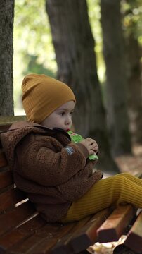 Sweet cute kid sitting on the bench in the park. Baby eats fruit puree from a pack and waving his foot. Vertical video