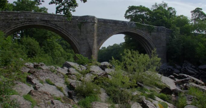Wide panning shot looking up to devils bridge from the river Lune with rocks in foreground at Kirkby Lonsdale, North Yorkshire