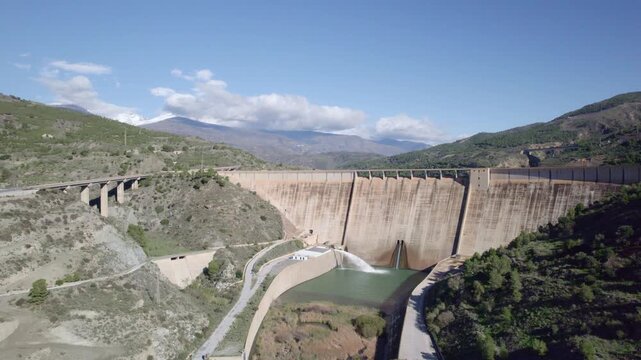 Gravity dam discharging water into the river. Howell bunger valve in operation. Hydroelectric power plant in operation. Panoramic aerial view over the dam and river. Renewable energies. Spain.