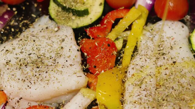Olive oil being drizzled over traybake of wild caught atlantic Cod fillets with mediterranean diet vegetables. Peppers, onions, courgette with fresh herbs. High quality healthy food videography.