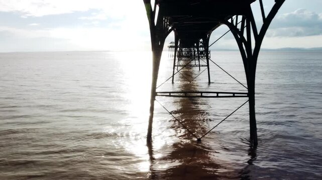 A drone shot sweeping under Clevedon Pier at sunset. It shows the pier leading out to sea. The sun reflects off the sea