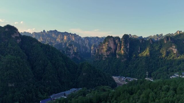 Long mountain walls and flat topped cliffs stretch above shadowed village in valley, aerial of Zhangjiajie China