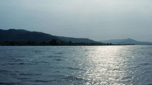 Rare slow aerial pullback reveals wide Vashishti River with reflective waters, vegetated banks, small boats, flanked by green hills in hazy evening light near Chiplun.