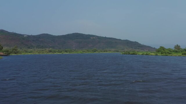 Slow drone glides above serene Vashishti River with calm blue waters bordered by lush green vegetation and trees, backed by hazy forested hills near Chiplun Maharashtra.