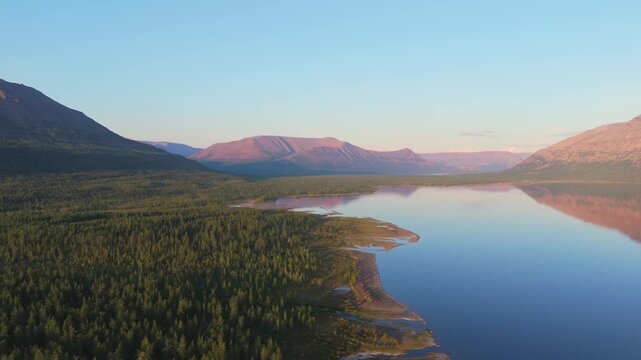 Warm golden hour light grazes Lake Lama shoreline on the Putorana Plateau, Russia. A gentle drone pass reveals evergreen forest, sandbar and lagoon, while rosy mountain reflection adds travel mood