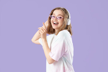 Fototapeta premium Cool young woman listening to music and dancing on lilac background