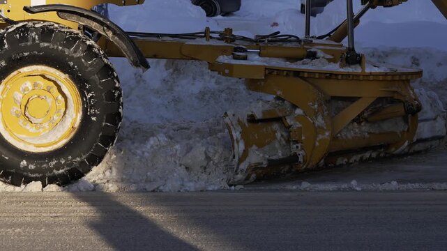 Close-up view of a yellow motor grader machine with a large steel blade removing compacted snow and ice from an urban roadway after a heavy winter storm, clearing the way for traffic