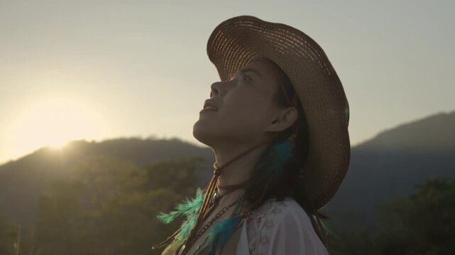 Close up of happy Asian woman looking up at the sky, admiring nature in boho style at sunset with mountain background and sun flare.