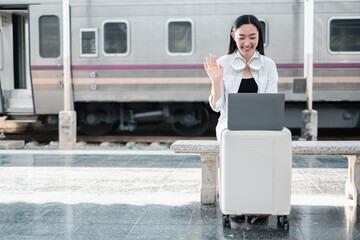 Smiling woman using laptop at train station, sitting on luggage, wearing headphones, waving during a video call.