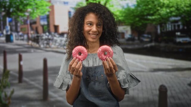 Teenage woman holding two pink donuts with both hands, smiling while wearing a denim apron and gingham blouse on a street by bicycles and trees; joyful sharing.