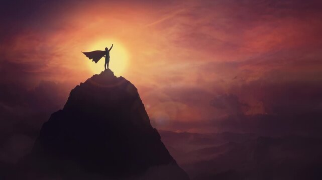 Conceptual sunset scene, superhero with cape standing brave on top of a mountain looks determined at horizon raising one hand up as a winning leader. Hero power and motivation, overcoming obstacles.