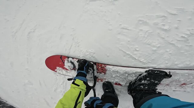 Snowboarder clipping into snowboard bindings at a ski resort. Board, gloved hands, and feet. Sports equipment. Lifestyle first-person view. Handheld