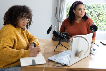 Female colleagues writing and recording podcast at wooden table with microphones, laptop