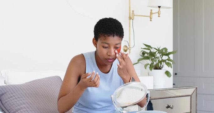 Checking mirror, adult African American woman sitting on bed applying red eye patch for skincare