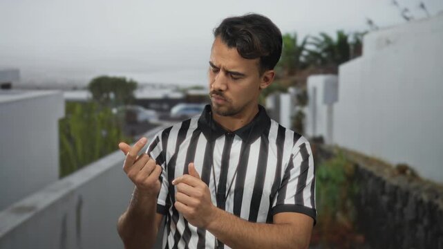 Young hispanic man referee counting outdoors intently staring at fingers wearing striped shirt amidst urban background suggesting focus and determination.