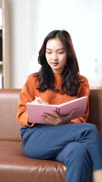 Young pensive asian woman sitting comfortably on a brown leather sofa in the living room, writing her thoughts, plans, or creative ideas in a personal pink notebook with a pen