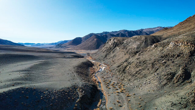 Sunlit Valley Winding Stream Through Arid Basin, Warm Tonal Contrasts Between Rocky Slopes And Blue Sky, Slender Watercourse Supporting Golden Grasses, Inviting Exploration And Contrast To Frozen