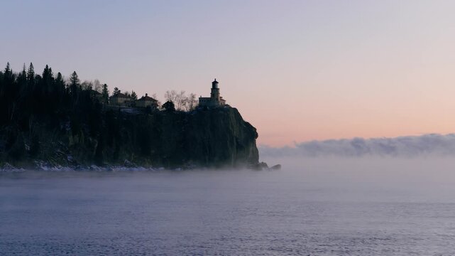 Split Rock Lighthouse emerges from heavy winter sea smoke and mist on a rugged cliffside during a subzero sunrise on Lake Superior.