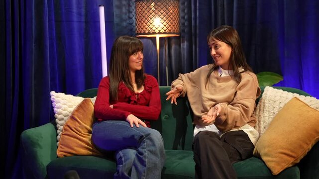 Two cheerful female content creators sitting on a comfortable couch and chatting while recording a video podcast or a talk show episode together in a professional streaming studio setup