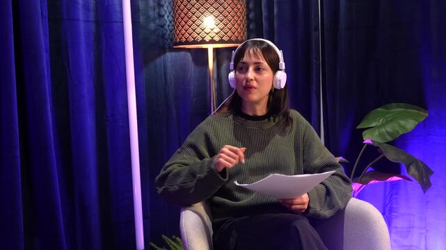 Expressive young woman wearing headphones and speaking passionately during a podcast interview, sitting in a cozy studio with blue curtains and warm lighting while actively gesturing