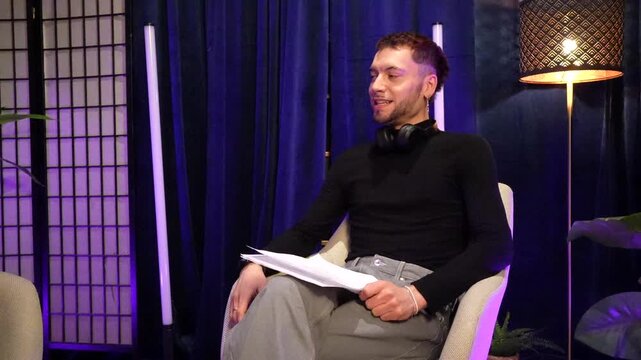 Young handsome man speaking and gesturing during an interview or podcast recording in a modern, stylishly lit studio, holding a script and engaging with an unseen person off camera