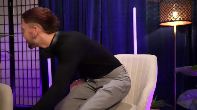 Smiling male content creator with headphones sitting in an armchair inside a recording studio, preparing to read questions from a script for a social media show or a live stream