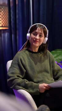 Pretty female influencer wearing headphones while recording a podcast or live stream for her social media channel, holding a script and smiling while listening to her guest's story