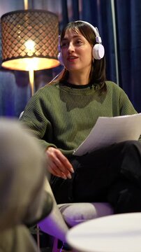 Caucasian woman wearing headphones speaking into a microphone while reading from a script, actively participating in a podcast recording session or a live broadcast in a professional studio