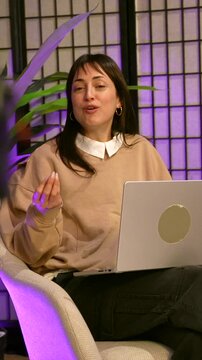 Young female content creator speaking and gesturing energetically while recording a video podcast or a vlog in a professional studio with purple ambient lighting and a laptop on her knees