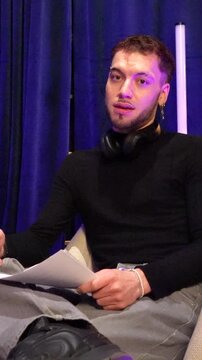 Stylish male podcaster with headphones and a script sitting in a studio, engaging with an audience during a live broadcast, expressing himself with dynamic gestures and facial expressions