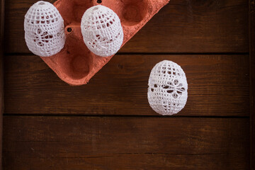 Handmade crochet Easter eggs in white lace covers placed on rustic wooden background with egg carton. Cozy craft concept, natural textures, copy space.