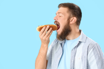 Portrait of handsome young man eating tasty hot dog on blue background