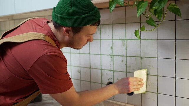 Concentrated male tiler in a green beanie and overalls carefully grouting white ceramic tile joints with green grout using a sponge during a kitchen renovation or remodeling project