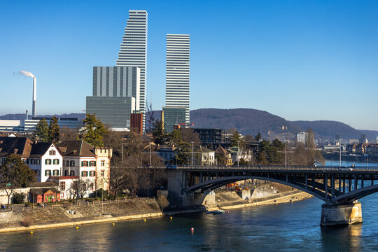 Modern cityscape of Basel, Switzerland, featuring the iconic Roche Towers and the Wettstein Bridge crossing the Rhine river under a clear blue sky.
