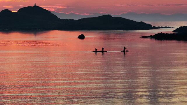 drone flight above sea at Ramberg beach, midnight sun in summer. 3 people on SUP board stand-up paddleboard enjoying sea and midnight sun in arctic Norway. Amazing nature, travel destination.