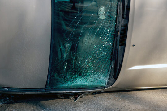 A tight close-up of a heavily shattered windshield of an overturned gold SUV after a traffic accident. The image captures the intricate spiderweb pattern of cracked safety glass and a small sticker in