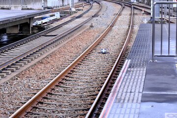 日本の鉄道駅のホームから眺める緩やかにカーブする線路。通勤や旅行の日常風景を象徴する空っぽのプラットホームと鉄路。輸送、交通インフラ、都市のライフスタイルをテーマにした、奥行きのある背景素材。 © taka