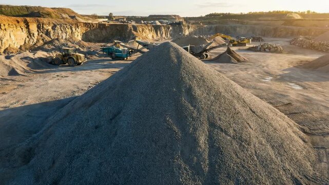 Geodesist using surveying instrument on gravel pile, showing quarry landscape and excavation vehicles at sunset for industrial site.