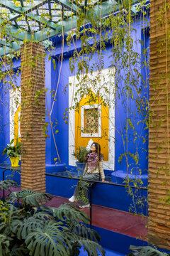 A woman sits on a blue ledge, framed by hanging vines, with a yellow window behind her. Jardin Majorelle,Marrakech,Morocco