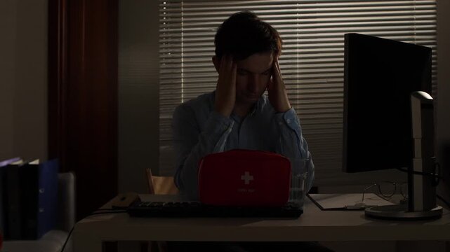 Stressed young man experiencing a severe headache or migraine while working late at his office desk, with a first aid kit nearby symbolizing a need for immediate relief