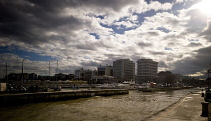 Porto urbano con barche ormeggiate, edifici costieri e cielo drammatico illuminato da raggi di sole
