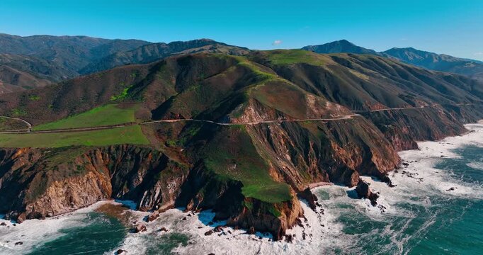 Aerial view of steep cliffs and Highway 1 in Big Sur. High angle shot of the coastal highway perched on a cliff edge above crashing ocean waves.