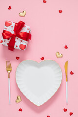 Romantic Valentine's Day vertical table setting with white heart shaped plate, golden cutlery, gift box with red ribbon and small hearts on pink background. Elegant flat lay concept for wedding or din