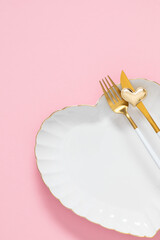 Elegant Valentine's Day table setting: White heart-shaped plate with gold rim, golden cutlery, and gold heart ornament on pink background. Romantic flat lay concept for wedding or anniversary.