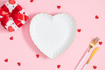 Elegant table setting for Valentine's Day: a white heart-shaped plate with a gold border, a gift box with a red ribbon, a gold fork and knife on a pink background. A flatlay wedding concept.